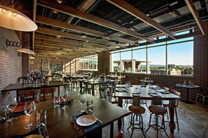 Un restaurante espacioso con techos altos, vigas a la vista y grandes ventanales con vistas a la ciudad.