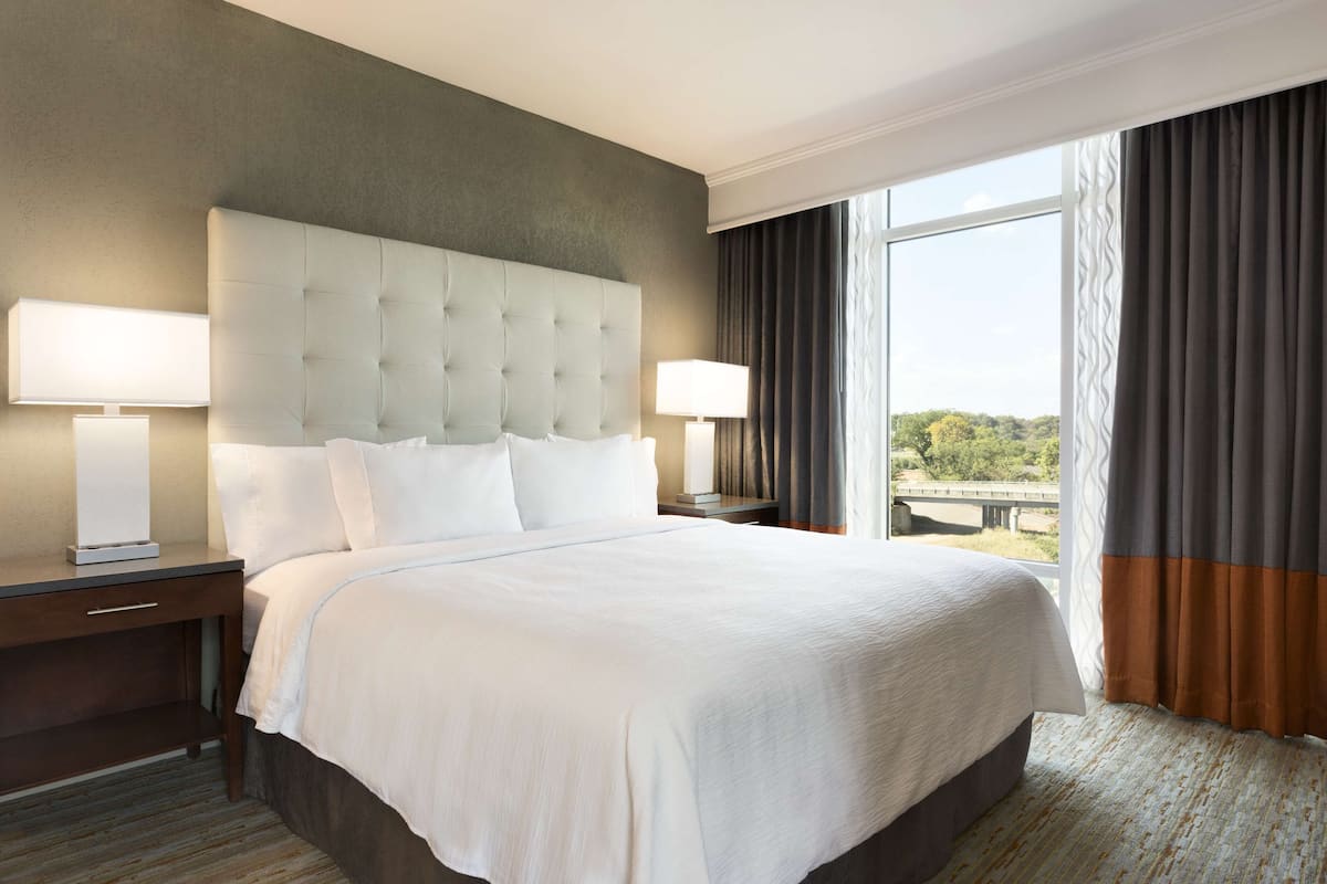 A large bed with a tufted headboard, two bedside tables with lamps, and a view of a bridge through a large window with curtains.