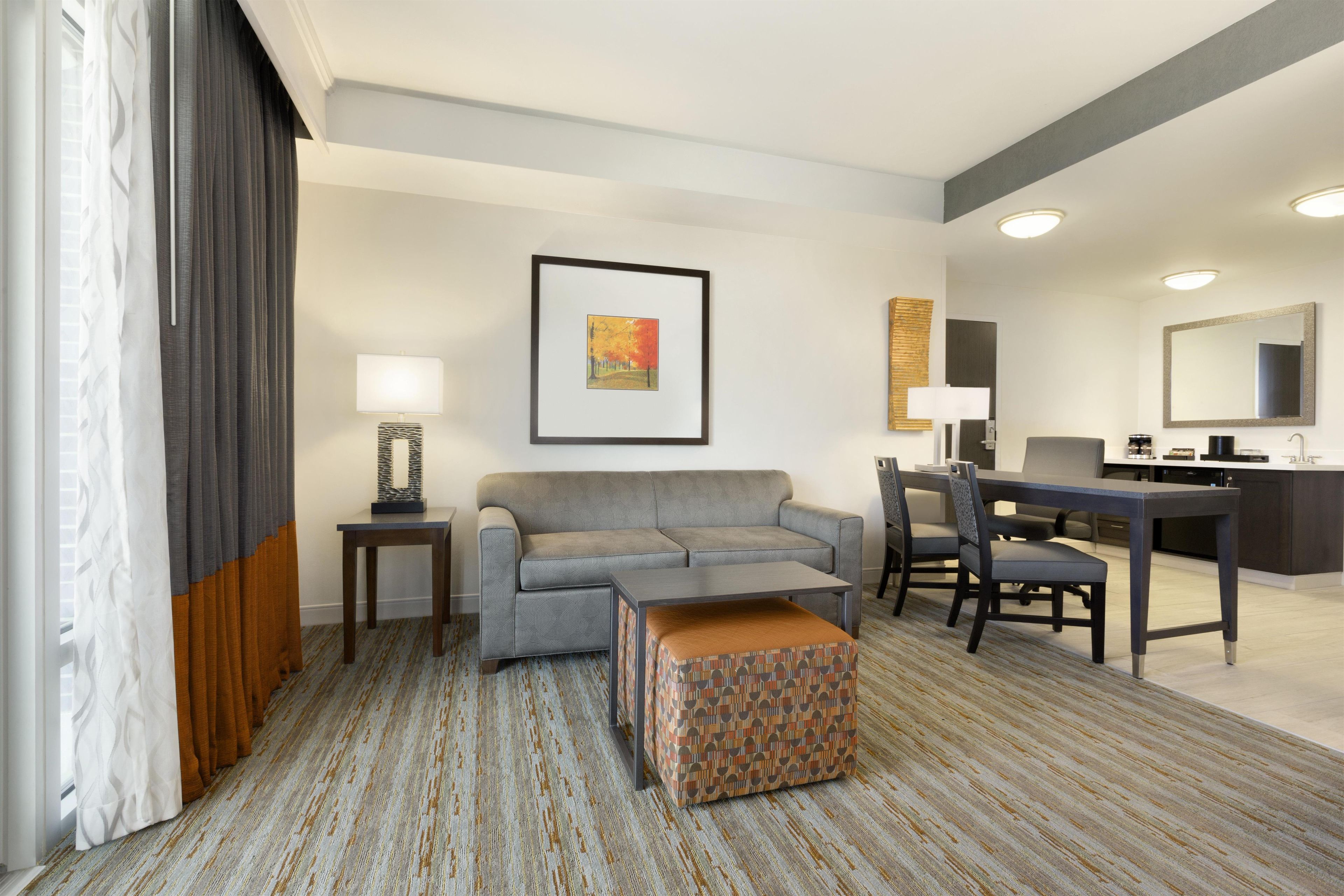 Une chambre d’hôtel avec un canapé gris, un petit pouf, une table à manger avec des chaises, un tableau encadré au mur et un grand miroir au-dessus du miroir.