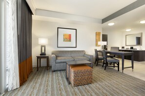 A hotel room with a grey sofa, a small ottoman, a dining table with chairs, a framed picture on the wall, and a large mirror above the mirror. - Embassy Suites by Hilton Greenville Downtown Riverplace (Greenville)