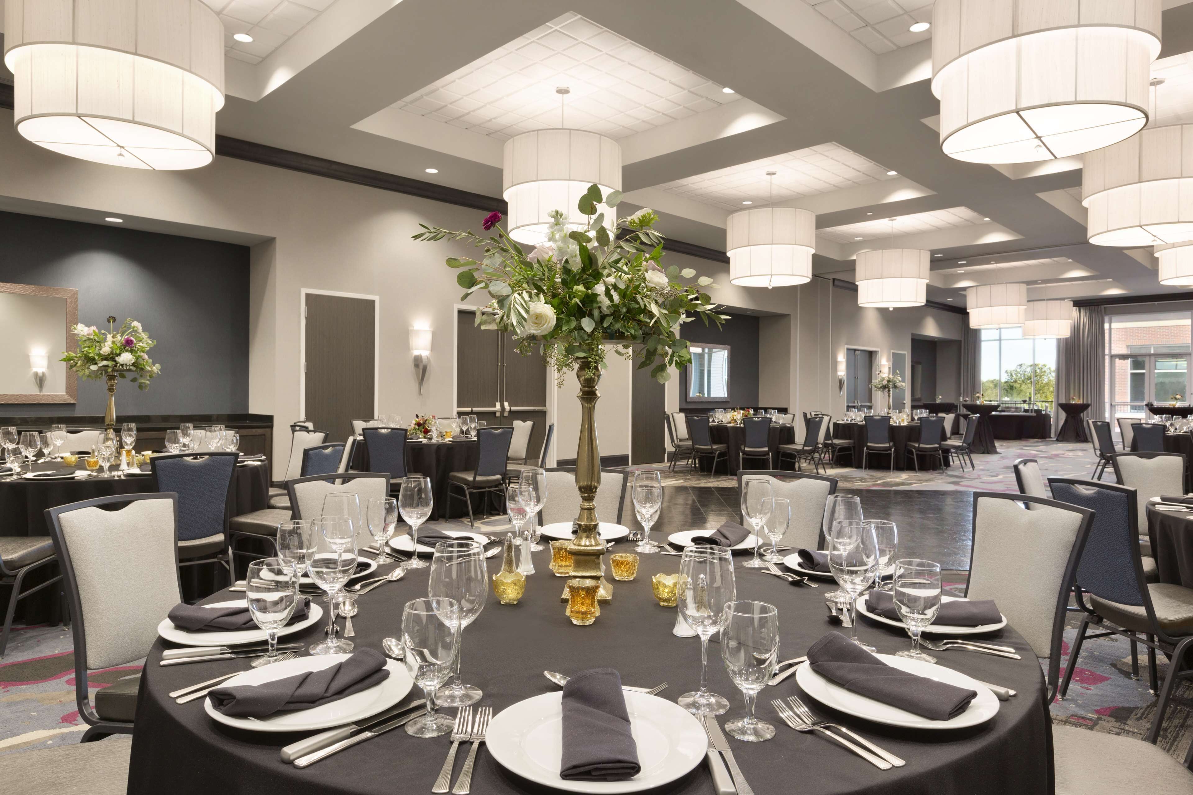A banquet hall with round tables set for dining, featuring white tablecloths, black napkins, and clear glassware.
