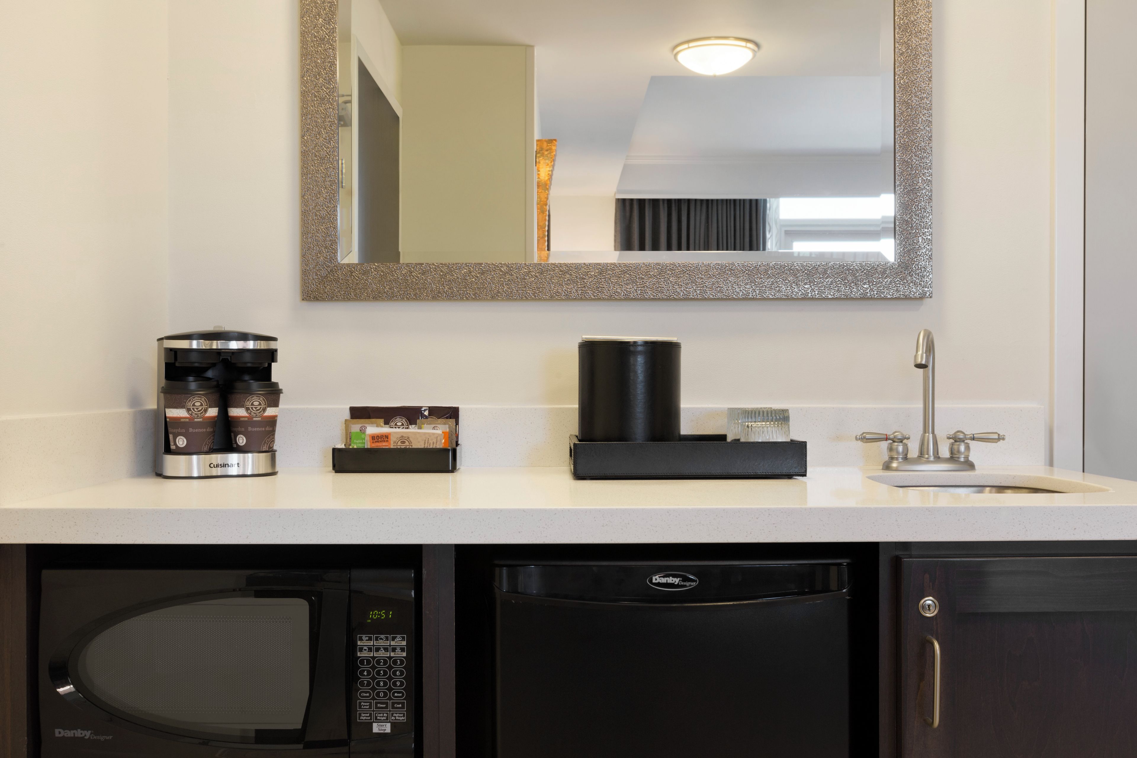 Un baño moderno con una encimera blanca, una cafetera, un microondas Danby Designer, un dispensador de jabón negro y un espejo con marco texturizado.