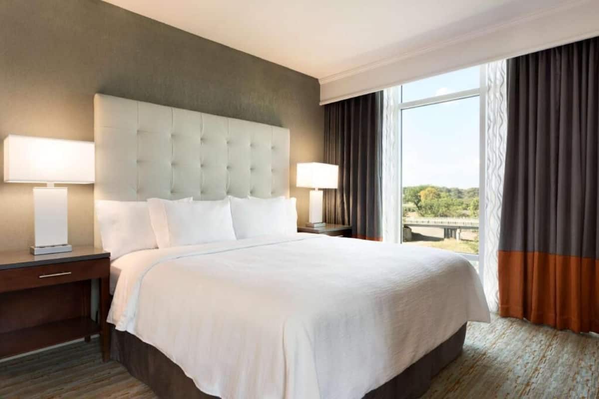 A hotel room with a large bed, two bedside tables, a window with curtains, and a view of greenery outside.