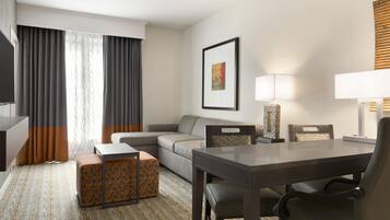A hotel room with a sofa, a dining table with chairs, a TV, a lamp, and a framed picture on the wall.