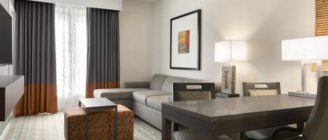 A hotel room with a sofa, a dining table with chairs, a TV, a lamp, and a framed picture on the wall.