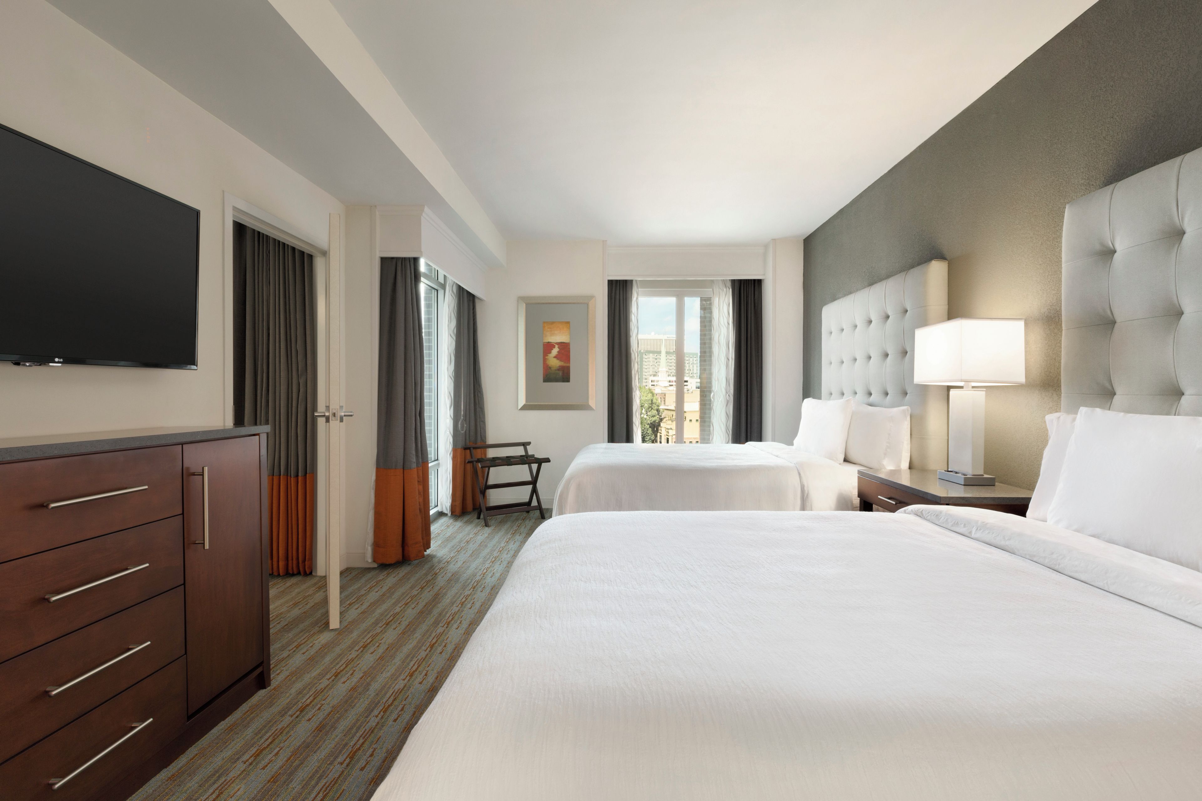 A hotel room with two beds, a TV, a desk, and a view of the city.