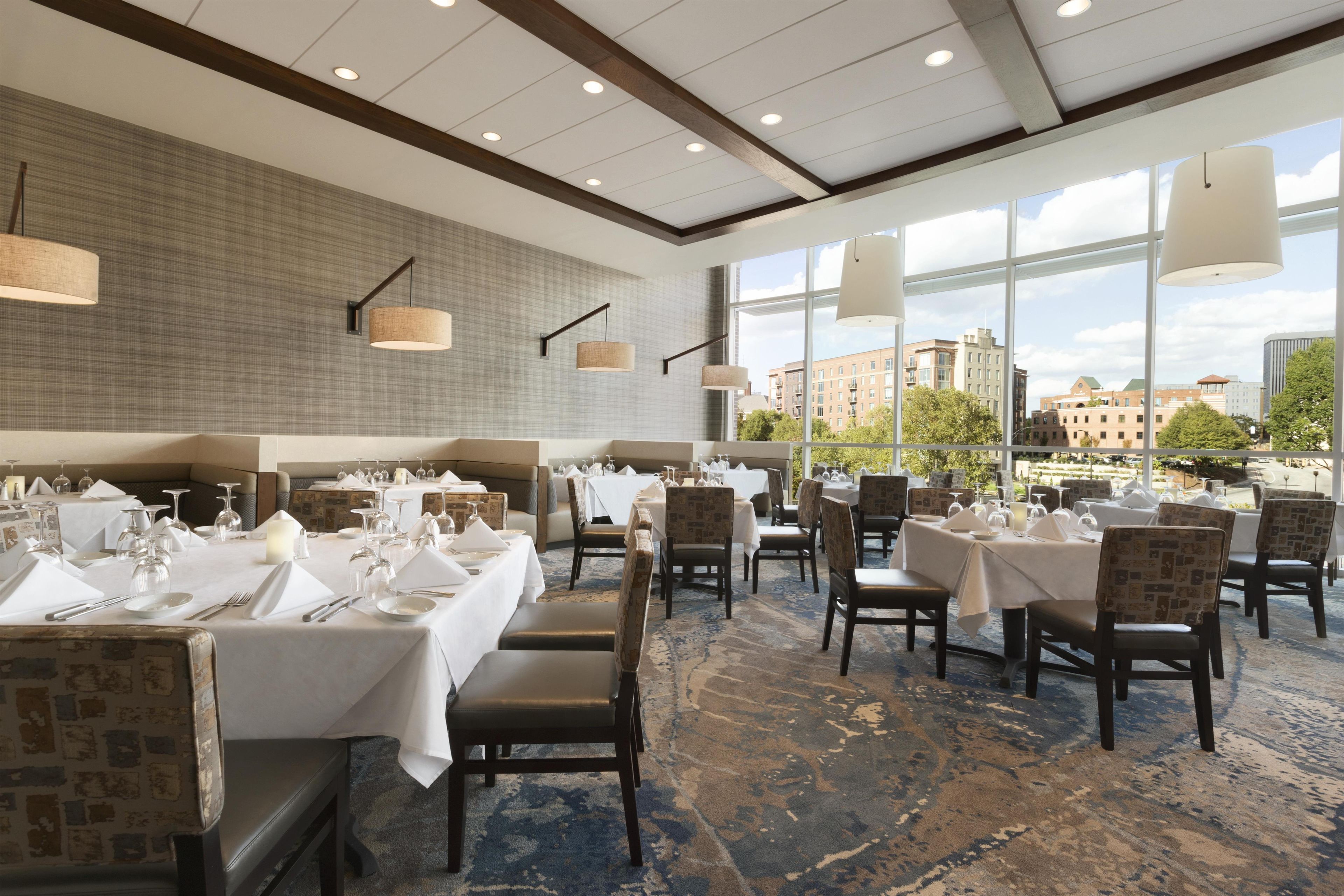A restaurant with multiple tables set for dining, large windows offering a view of buildings and trees, and a patterned carpet.