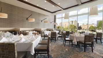 A restaurant with multiple tables set for dining, large windows offering a view of buildings and trees, and a patterned carpet.