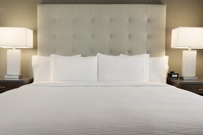A neatly made bed with a tufted headboard, two bedside lamps, and a digital clock on a bedside table. - Embassy Suites by Hilton Greenville Downtown Riverplace (Greenville)