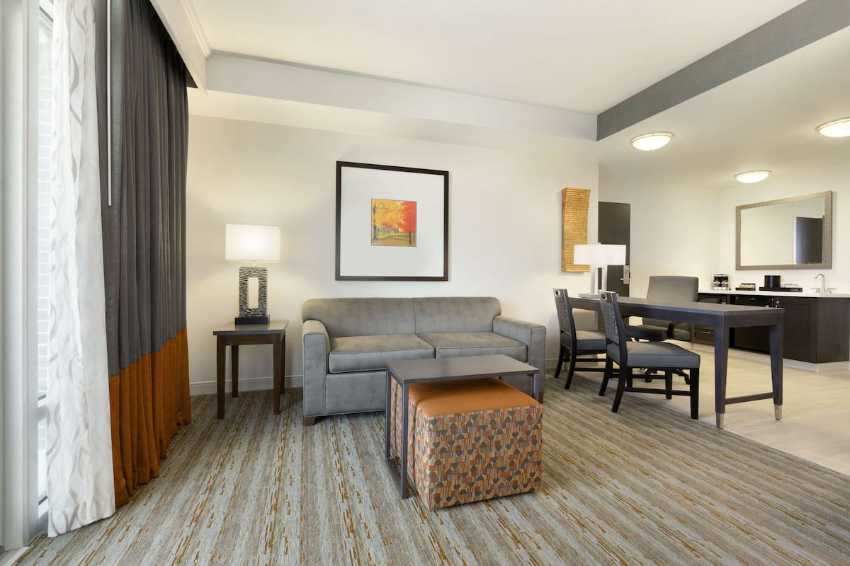 A modern hotel room with a grey sofa, a small table, a patterned ottoman, a desk with a chair, a large mirror, and a framed picture on the wall.