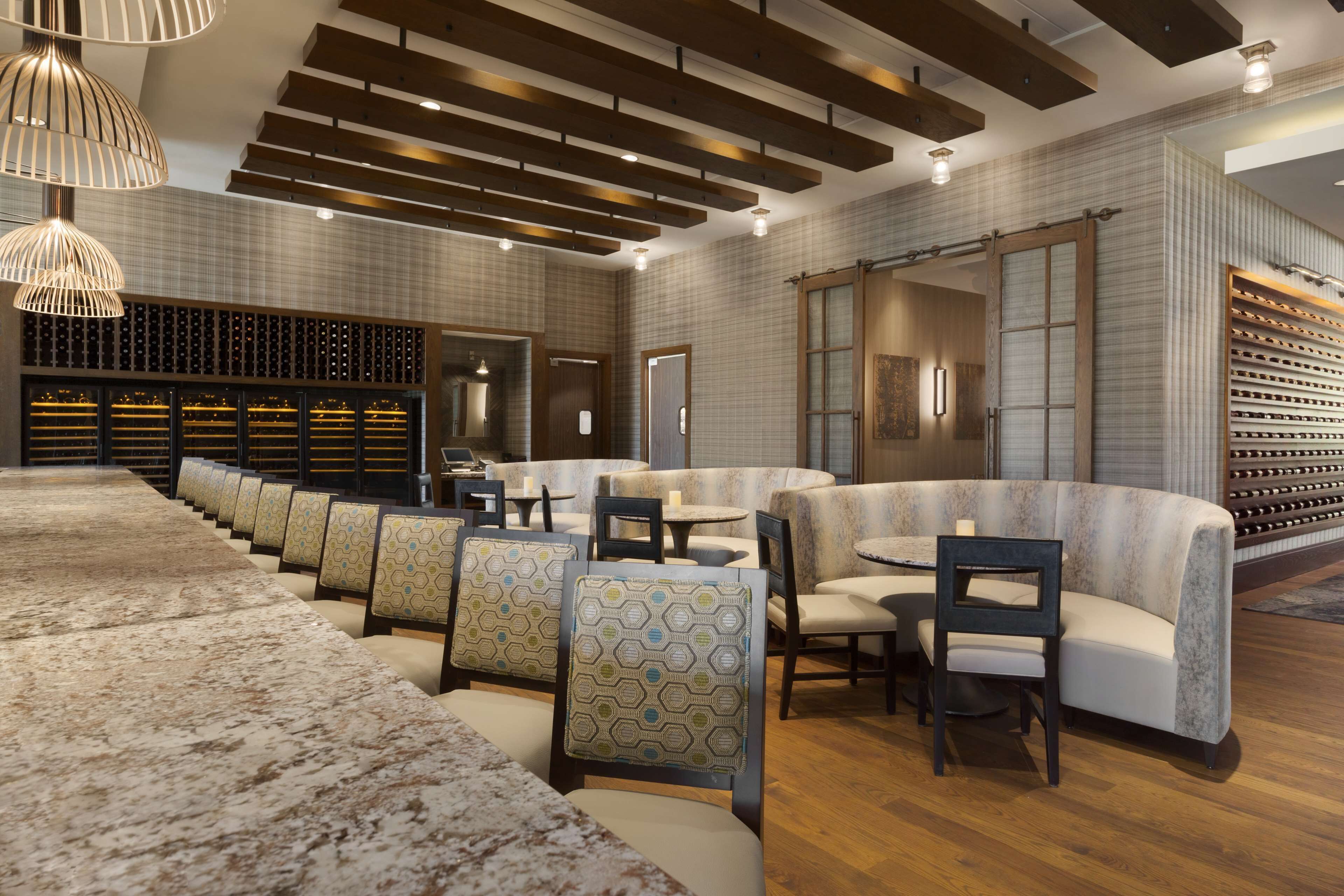 A modern restaurant with a long dining table, cushioned seating, pendant lighting, and a wine rack.