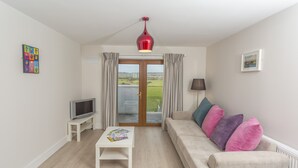 A living room with a sofa, a coffee table, a TV, and a view of the outdoors through a glass door. - Garryvoe Beach Homes (Garryvoe)