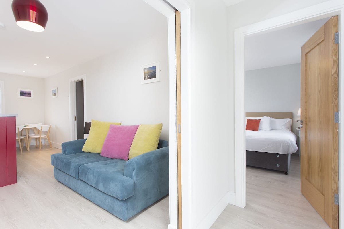 A modern, well-lit room with a blue sofa, a wooden door, and a bed with white bedding.