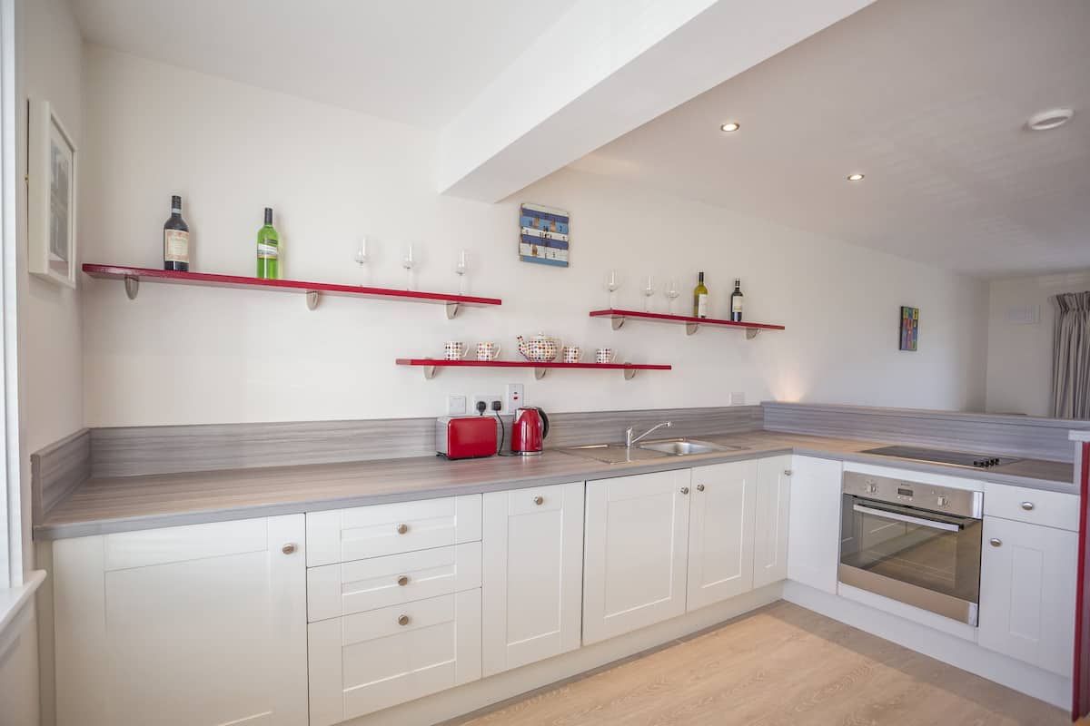 A modern kitchen with white cabinets, a built-in oven, and red shelves holding wine bottles and glasses.