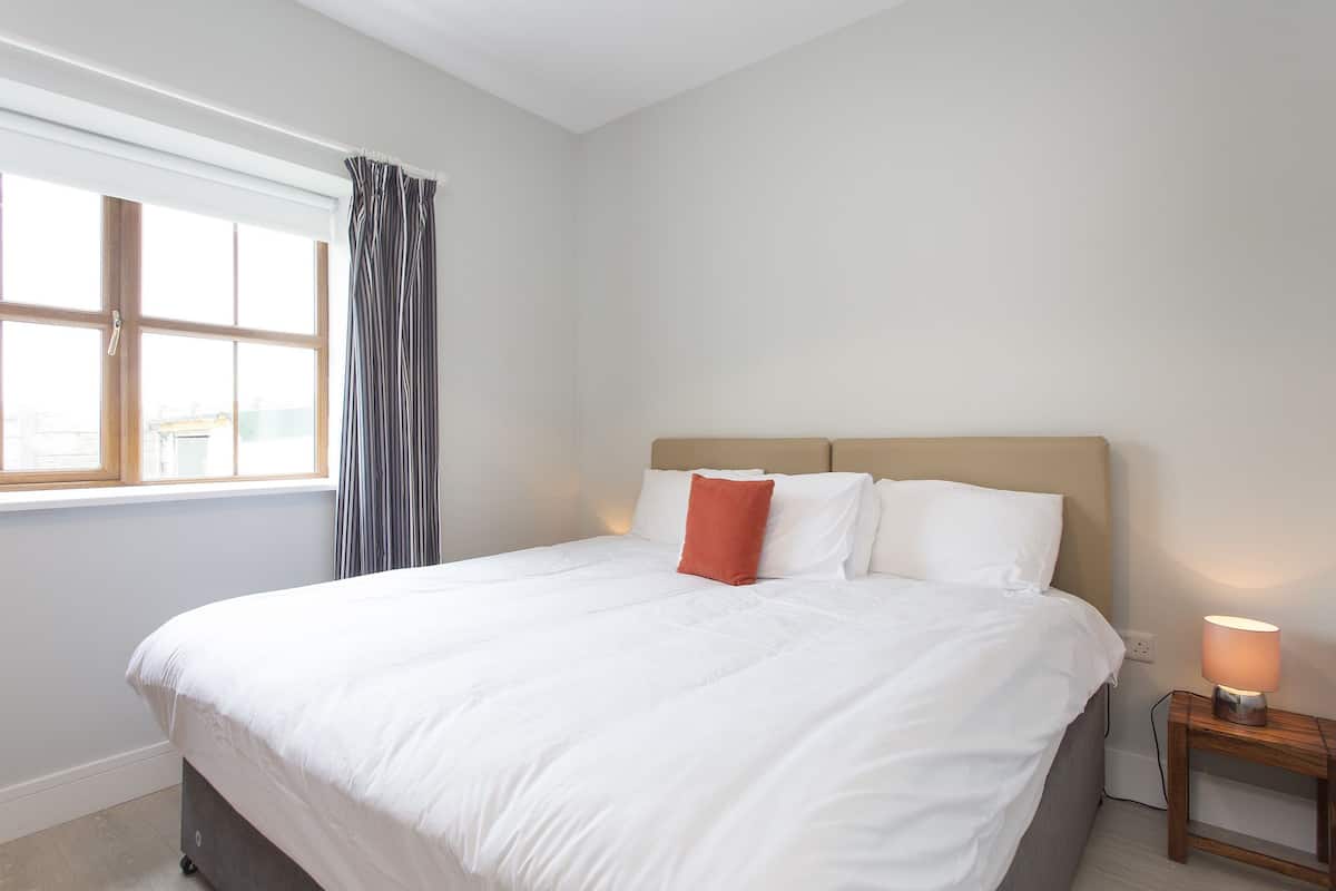 A neatly made bed with white linens and a single red pillow, a bedside table with a lamp, and a window with curtains.