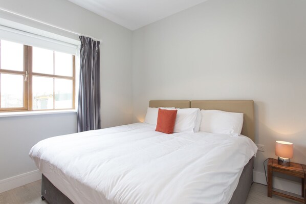 A neatly made bed with white linens and a single red pillow, a bedside table with a lamp, and a window with curtains.