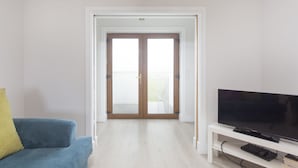 A living room with a blue sofa, a television on a white stand, and a door leading to an outdoor area. - Garryvoe Beach Homes (Garryvoe)