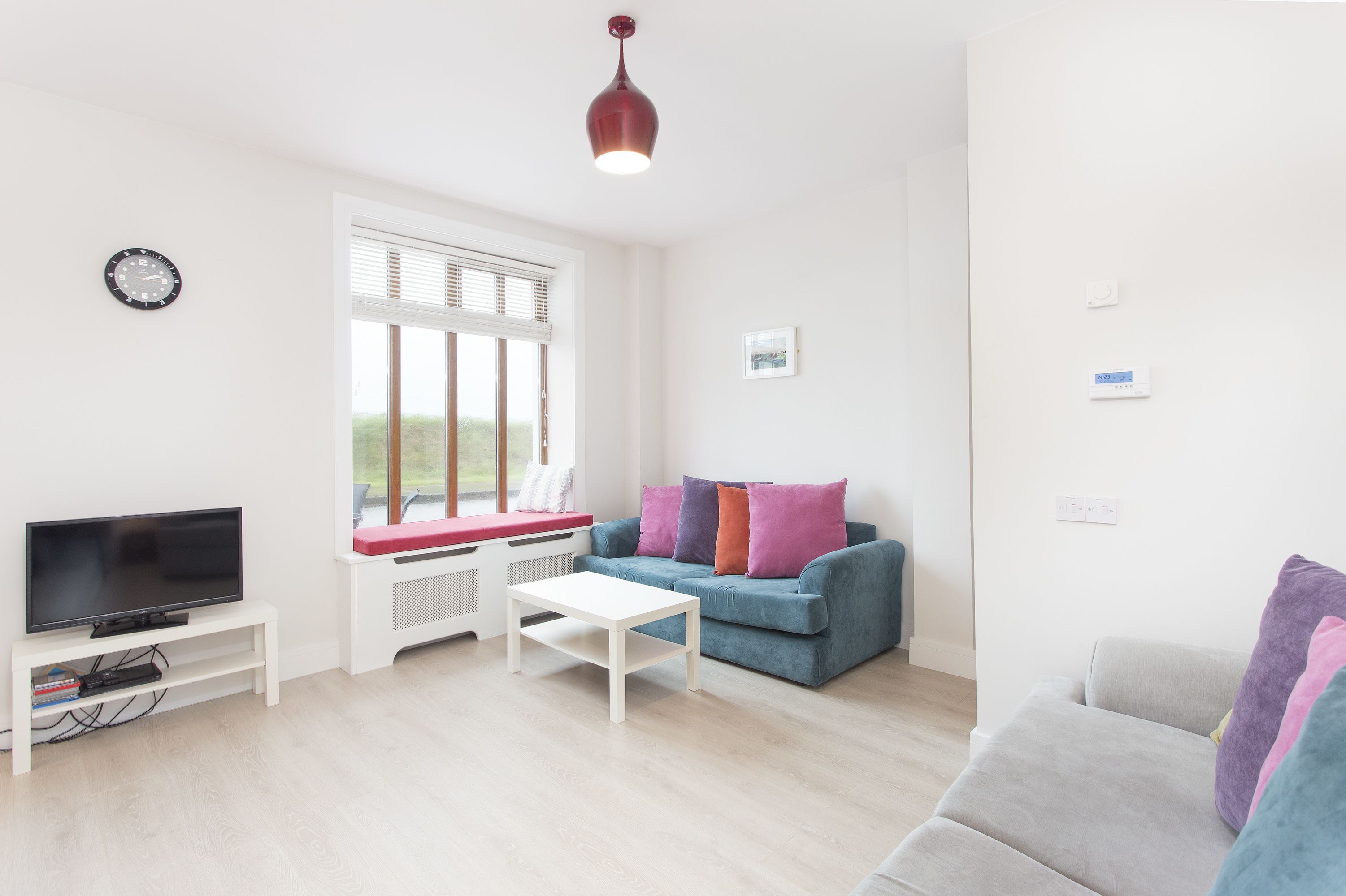 A modern living room with a flat-screen TV on a white stand, a white coffee table, a blue sofa with colorful cushions, and a window with blinds.