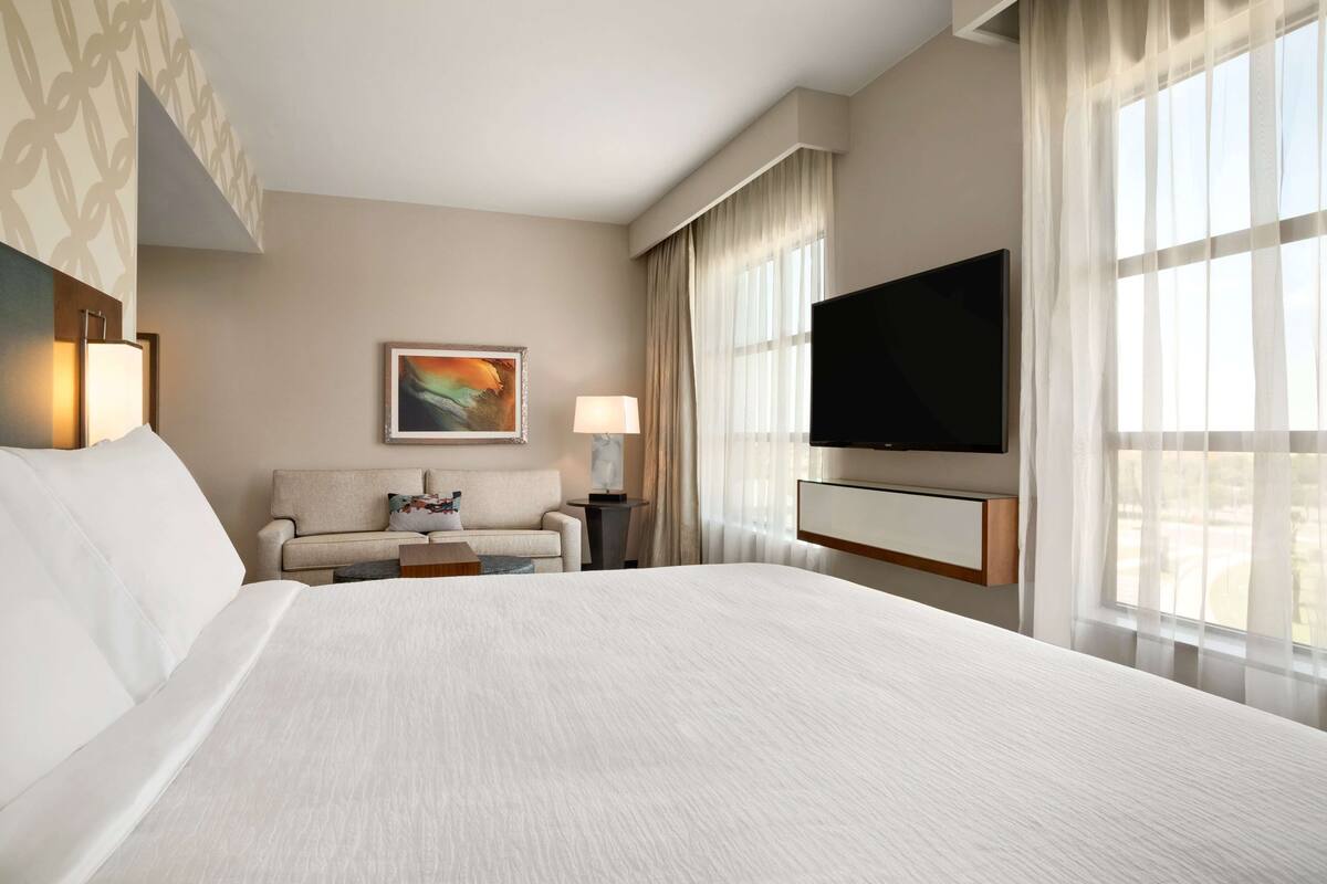 Une chambre d’hôtel avec un grand lit, un canapé, un téléviseur fixé au mur et une fenêtre ornée de rideaux légers.