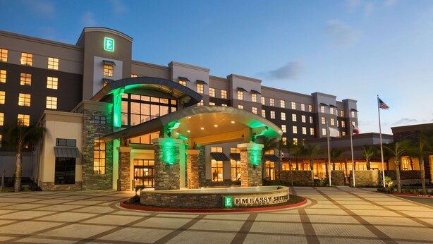 A hotel entrance with a covered waiting area, a flag, and a sign indicating it's an Embassy Suites. - Embassy Suites by Hilton McAllen Convention Center (McAllen)