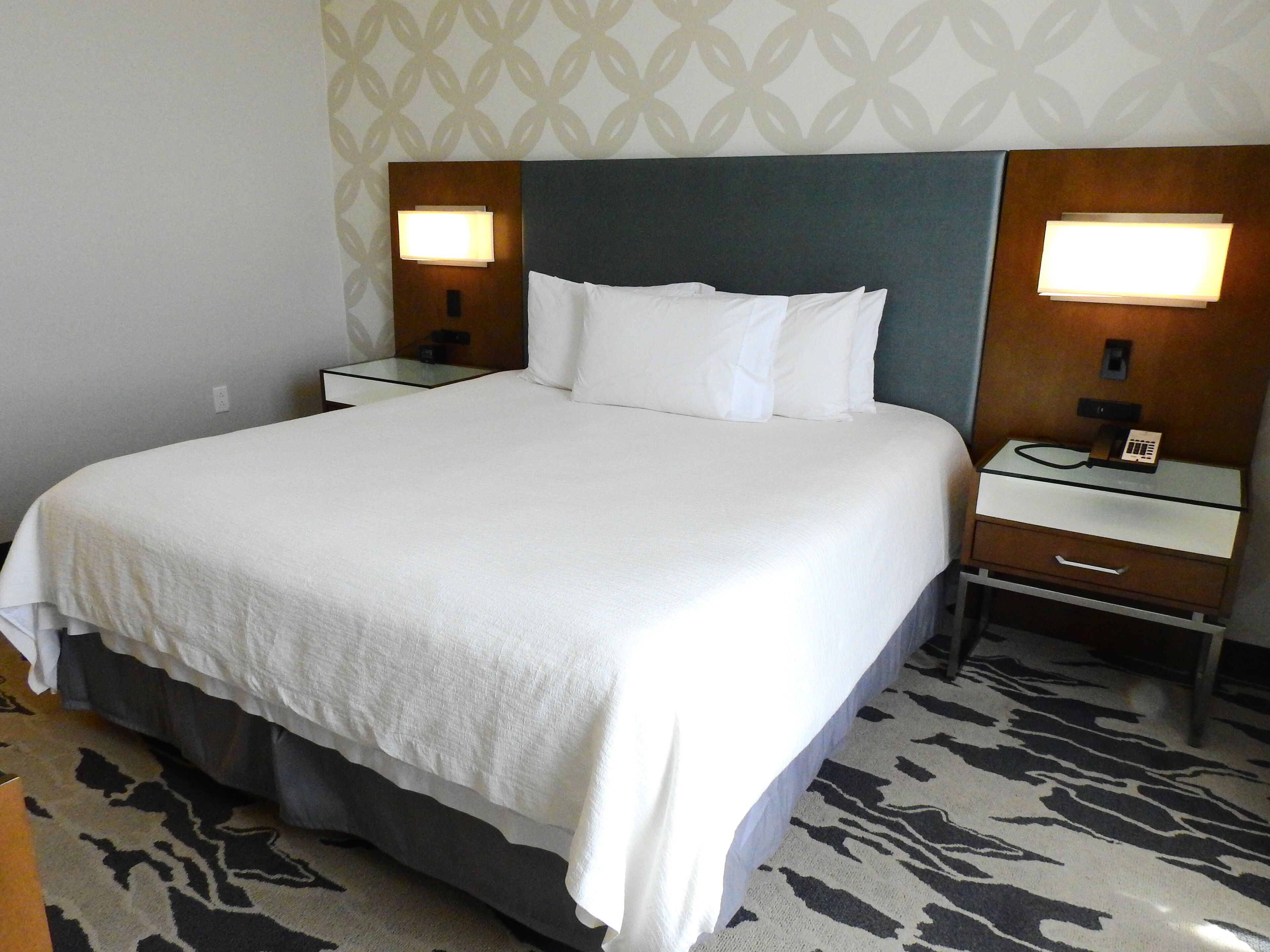 Una habitación de hotel con una cama grande, dos mesitas de noche con lámparas, una mesita con un teléfono y una alfombra estampada.