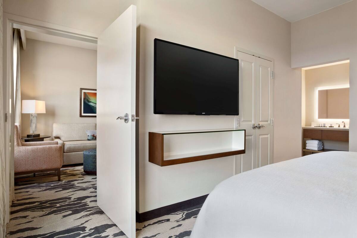 Une chambre d’hôtel avec un lit, un téléviseur fixé au mur, un canapé, une lampe et un tableau accroché au mur.