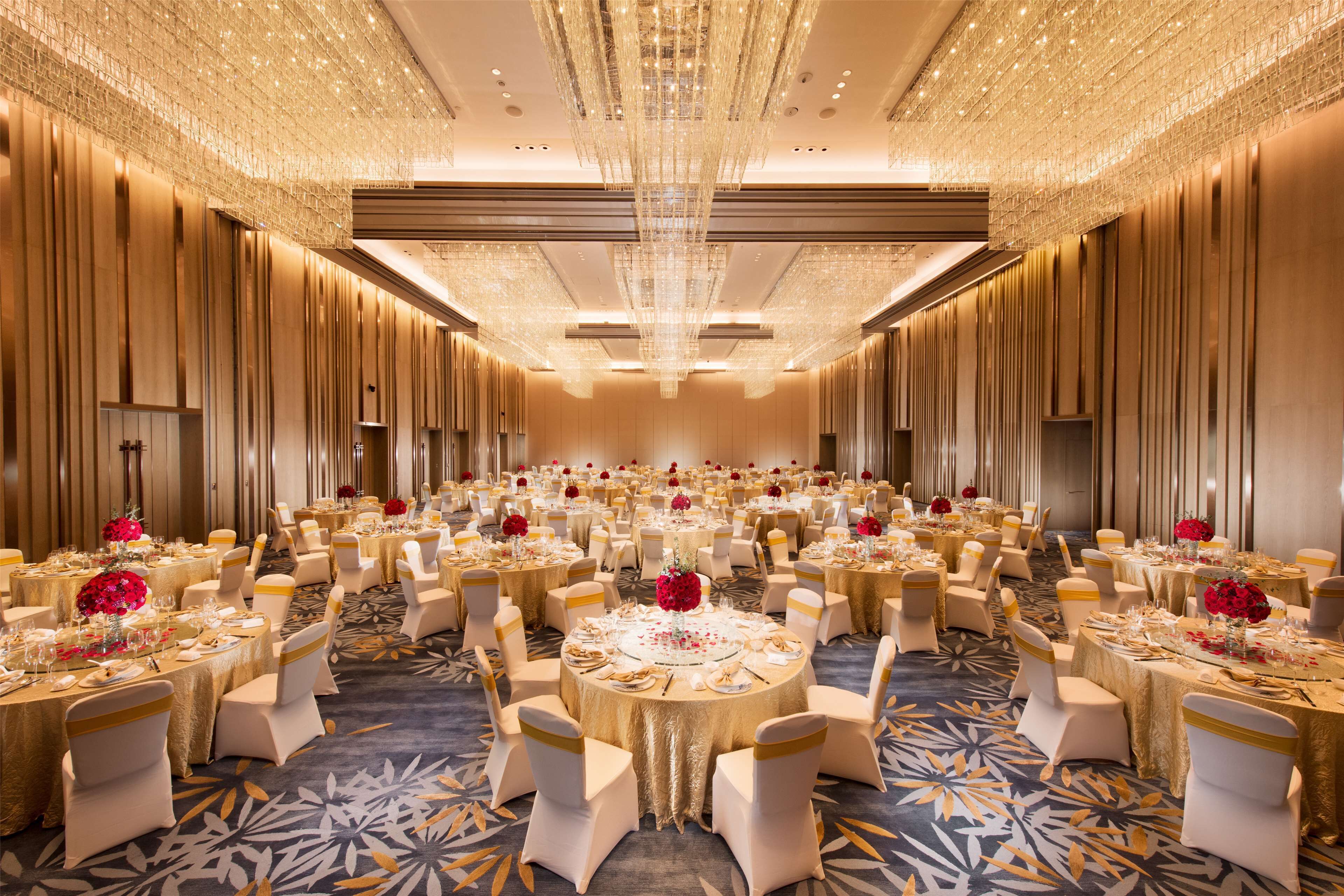 A banquet hall with round tables, white chairs, and red floral centerpieces.