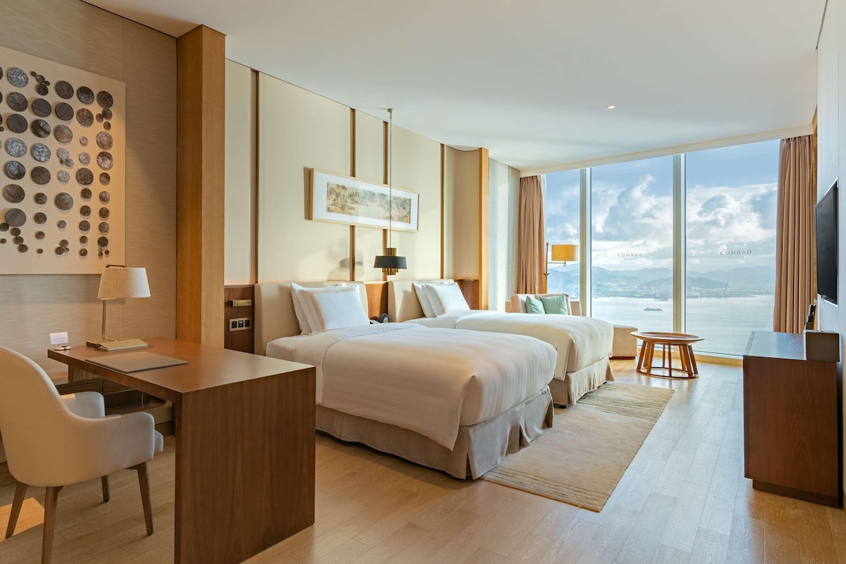 A hotel room with two beds, a desk, a chair, a TV, a large window with a view of the sea, and a wall with circular decorations.
