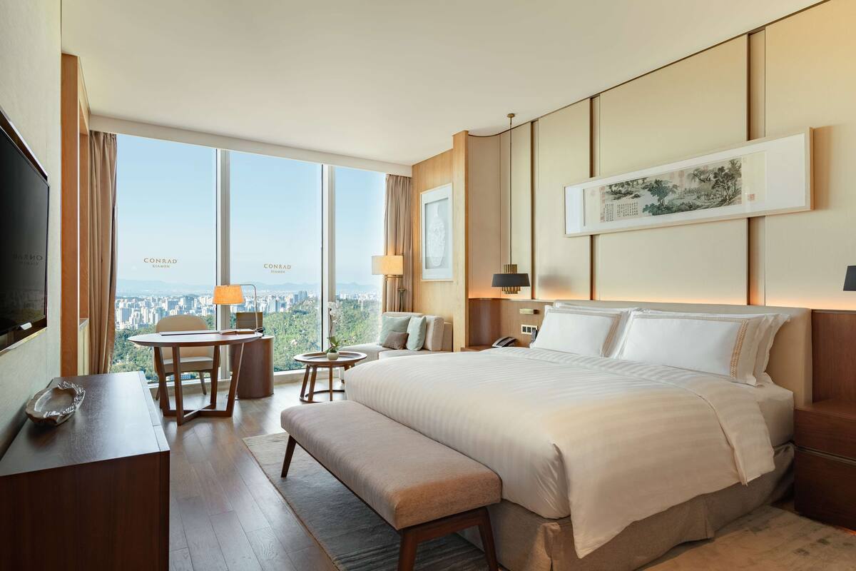 A hotel room with a large bed, a desk with a chair, a small table with a lamp, a bench, and a large window offering a city view.