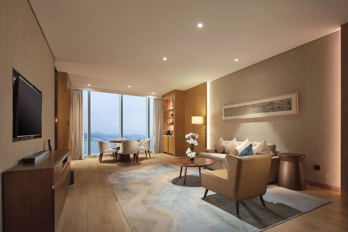 A modern hotel room with a flat-screen TV, a sofa, a coffee table, and a dining area with chairs.