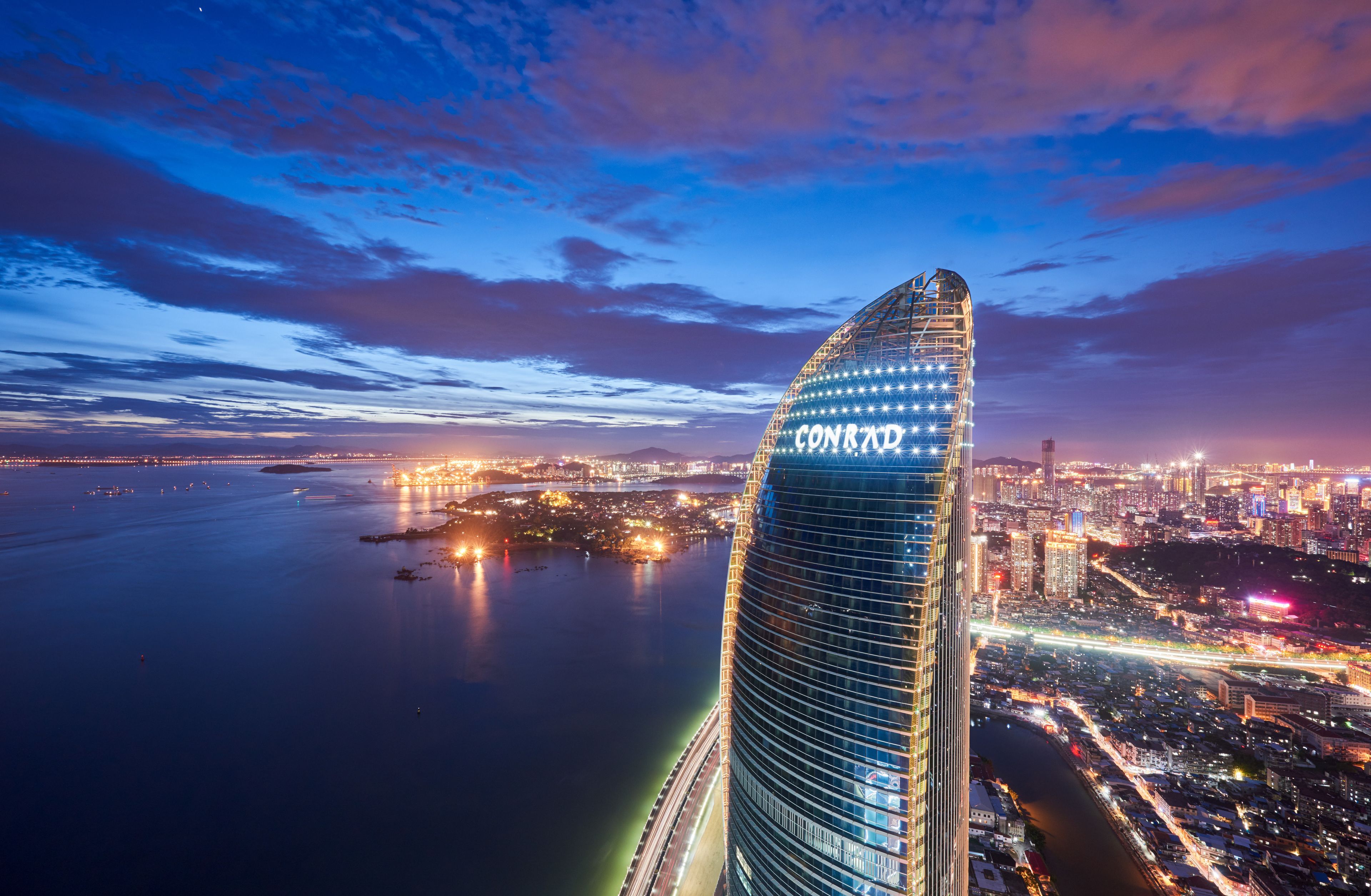 A modern skyscraper with a curved design, located near a body of water with city lights in the background.