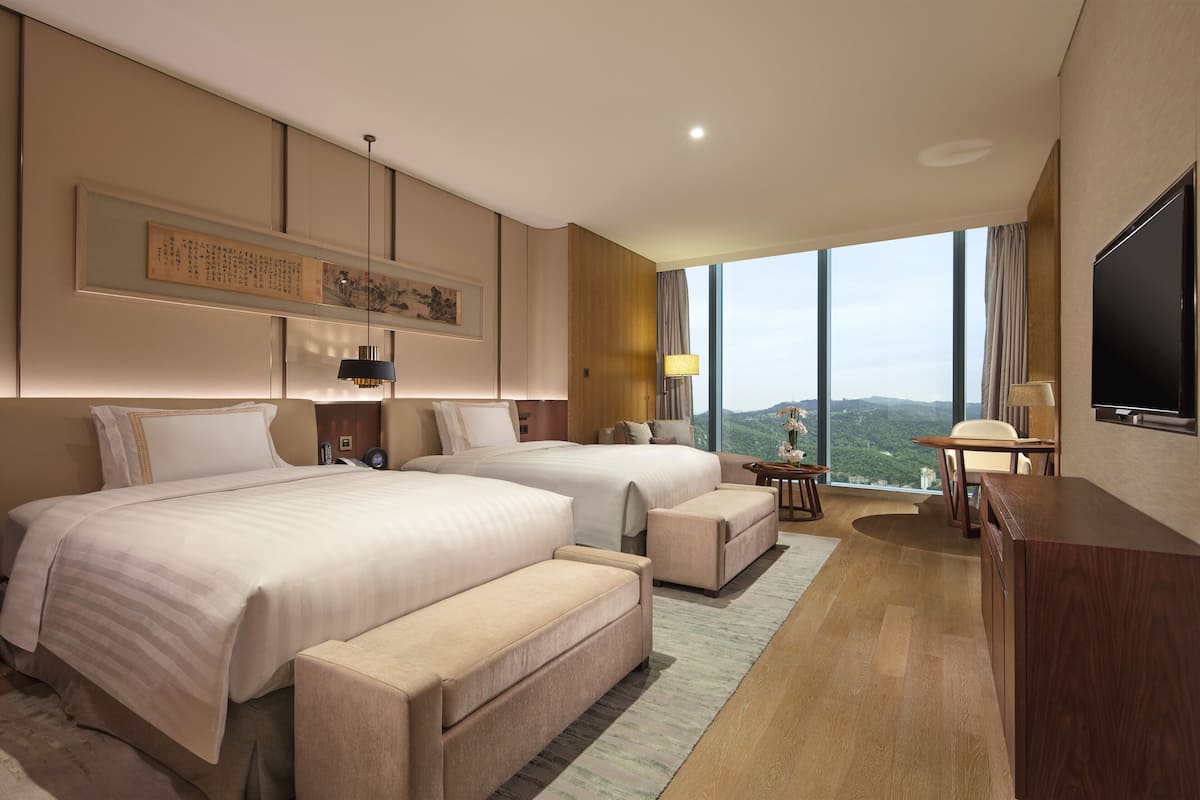 A modern hotel room with two beds, a desk, a TV, and a large window offering a city view.