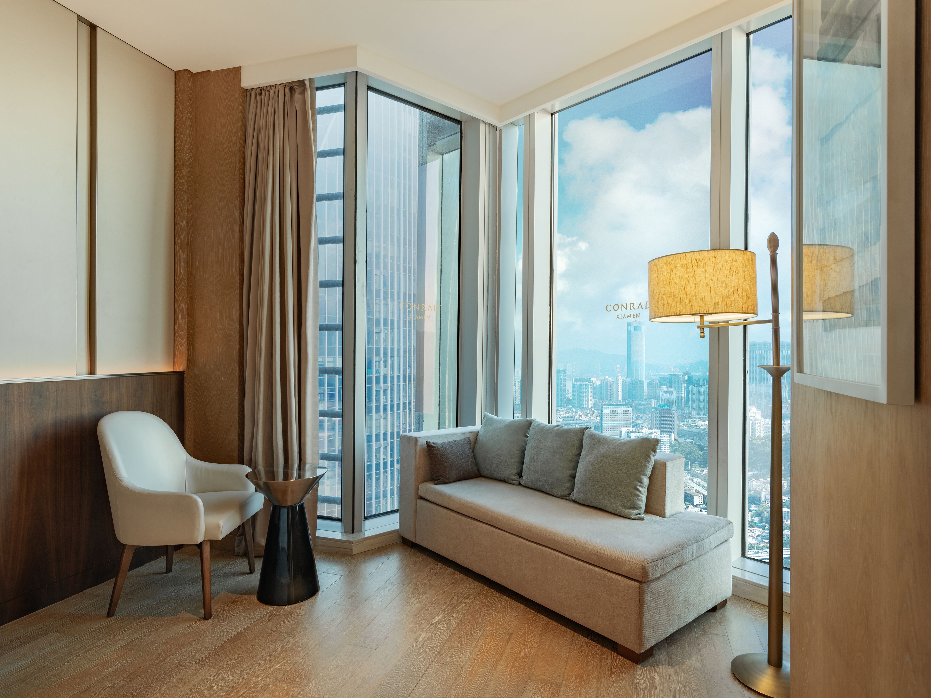 A modern hotel room with a large window offering a city view, a sofa, a chair, and a floor lamp.