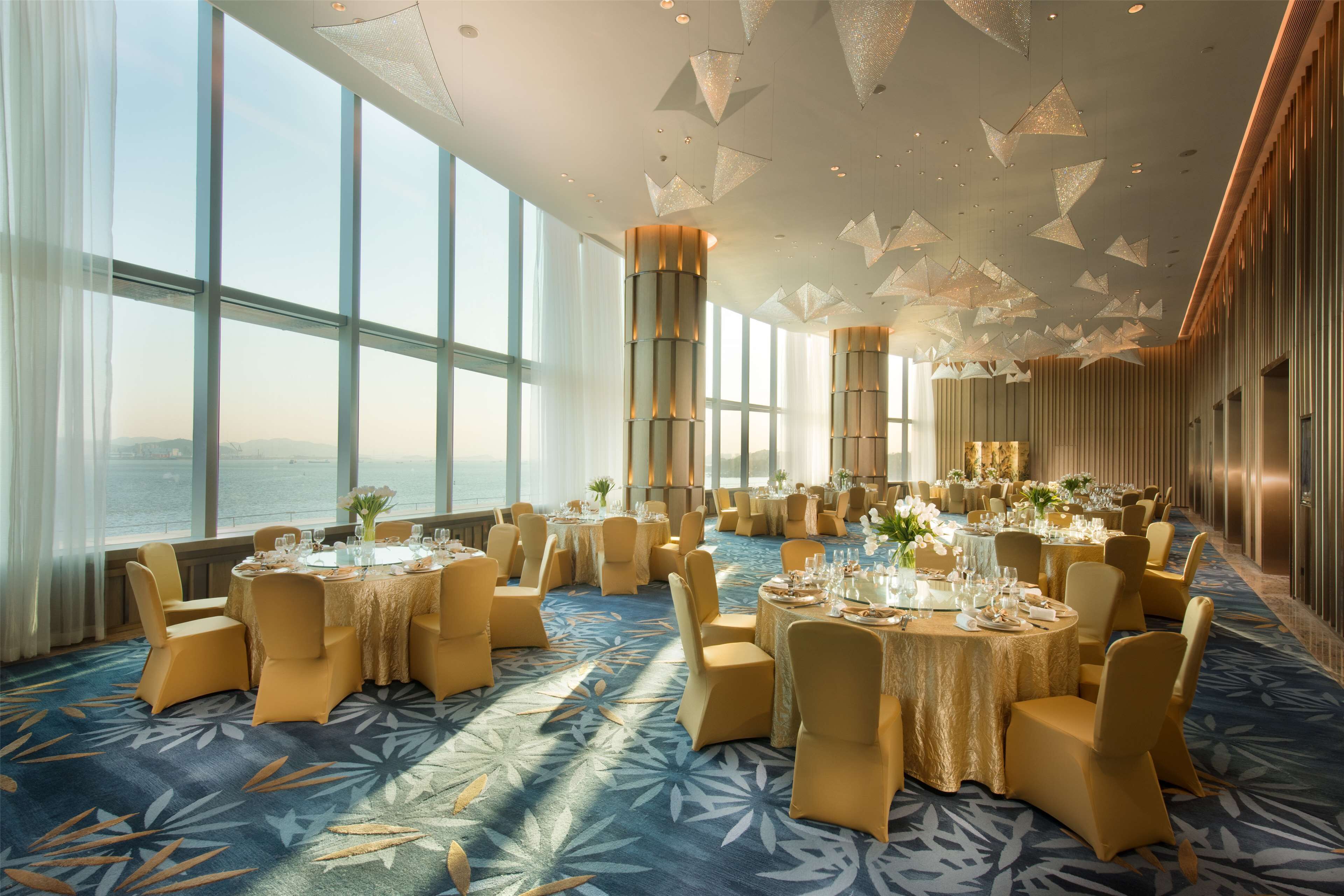 Une salle de banquet spacieuse, avec des tables rondes, des chaises dorées et une vue sur la mer grâce à de grandes fenêtres.