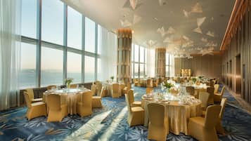 Une salle de banquet spacieuse, avec des tables rondes, des chaises dorées et une vue sur la mer grâce à de grandes fenêtres.