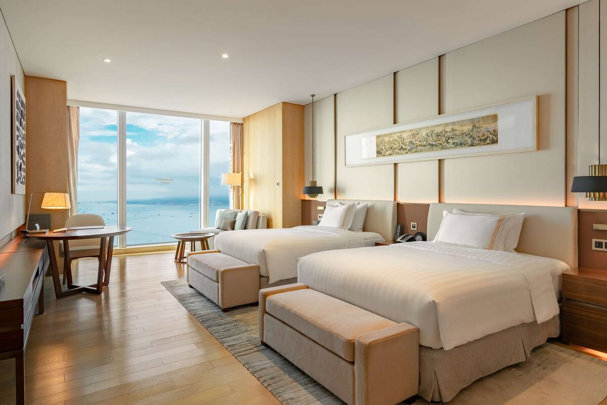 A hotel room with two beds, a desk, a chair, a laptop, and a view of the sea.