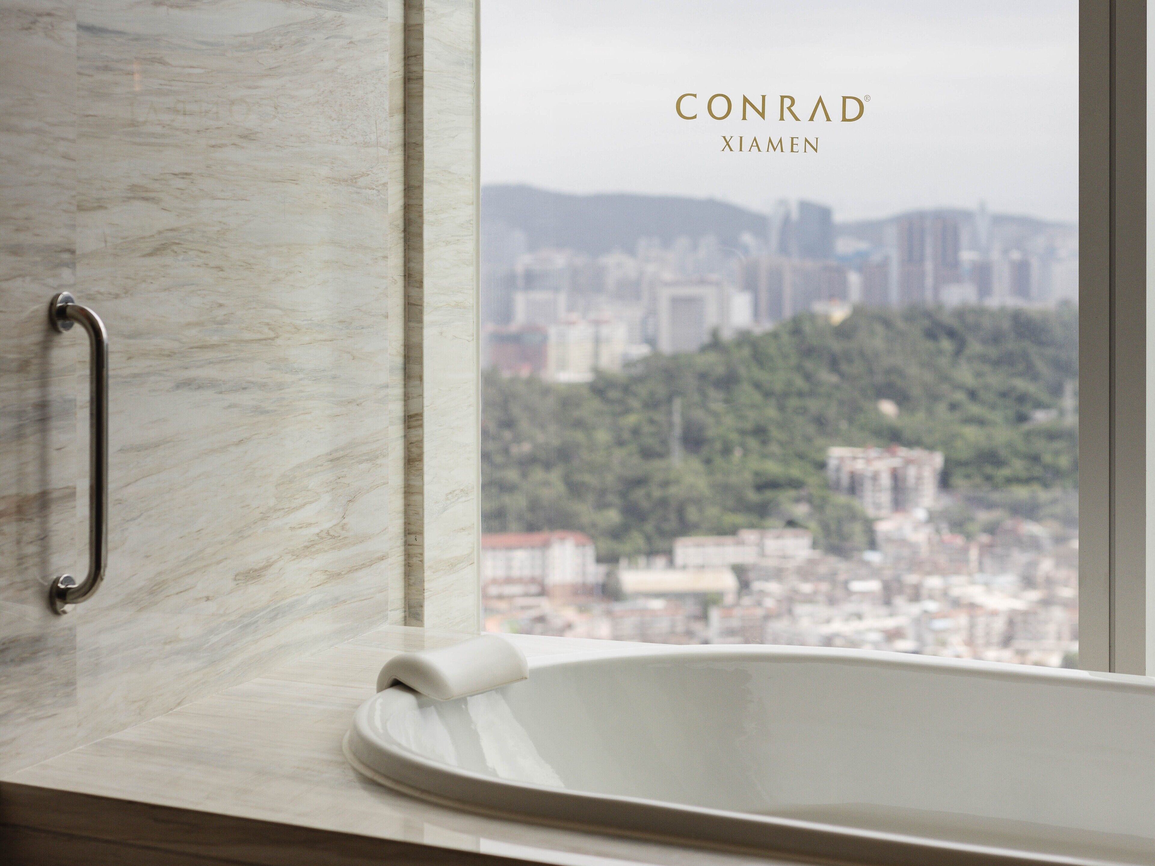 A bathroom with a bathtub, a marble wall, and a view of a cityscape through a window.