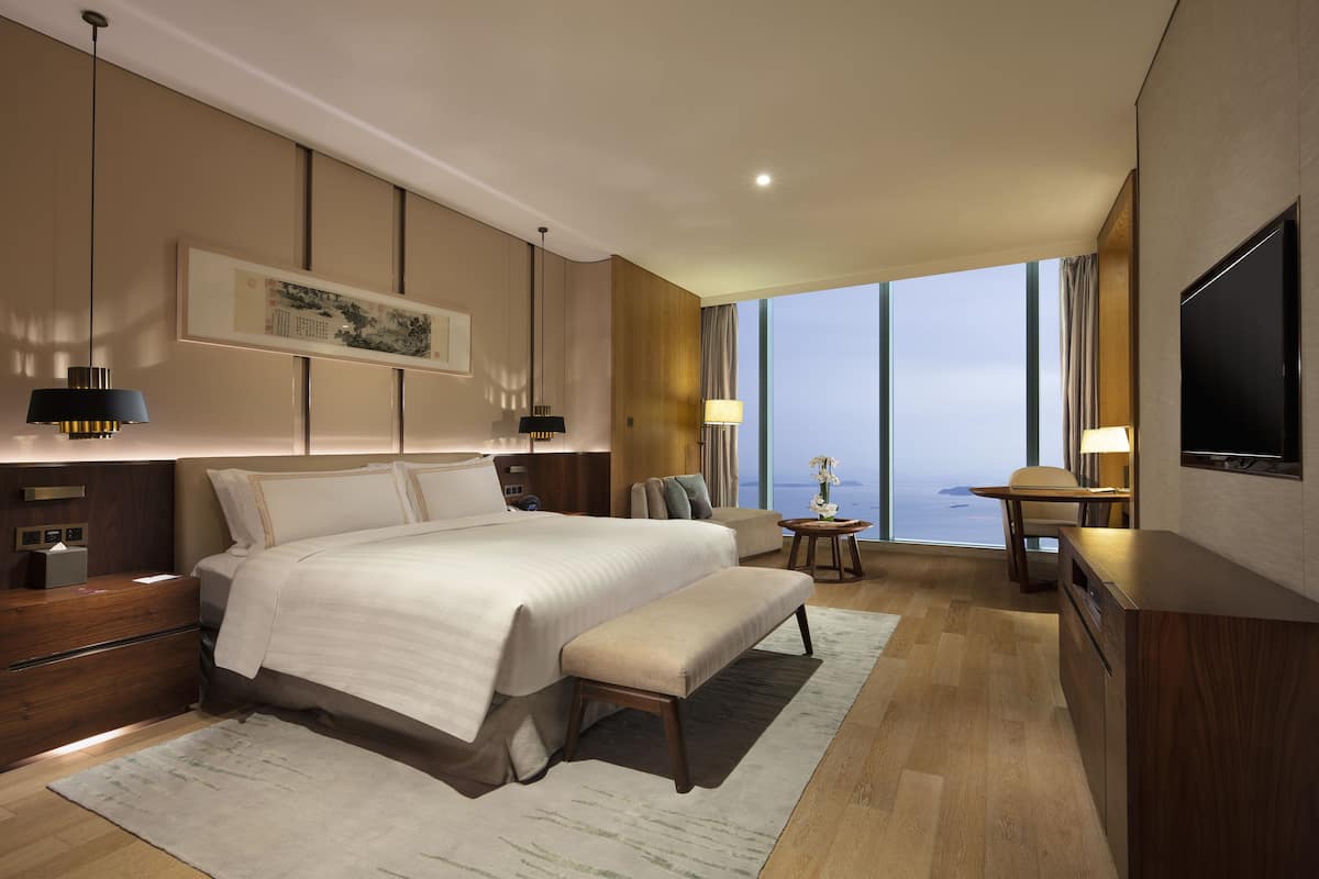 A modern hotel room with a large bed, bedside tables, a TV, and a view of the city.