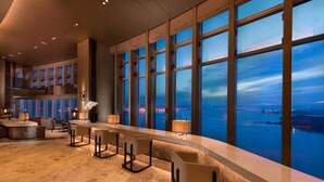 A modern bar area with a long counter, several bar stools, and large windows offering a view of the cityscape at night. - Conrad Xiamen (Xiamen)
