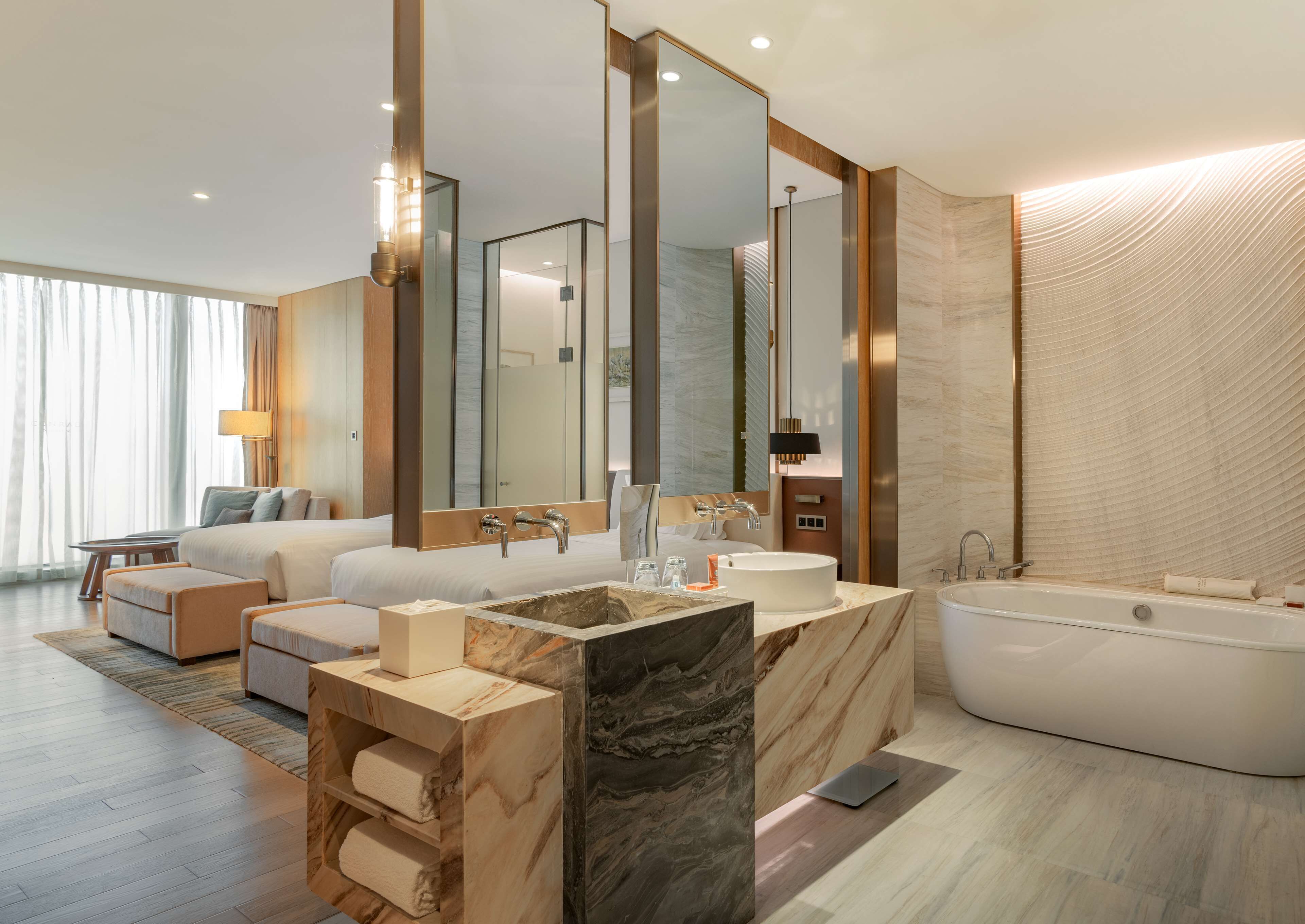 A modern hotel bathroom with a large mirror, a bathtub, a vanity with a marble countertop, and a large window with curtains.