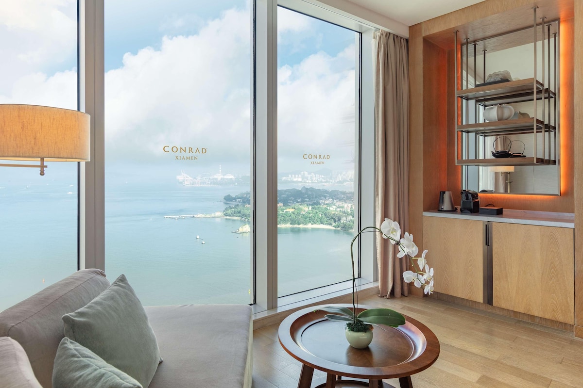 A modern hotel room with a large window offering a city and water view, a cozy seating area, and a small kitchen with a built-in shelf.