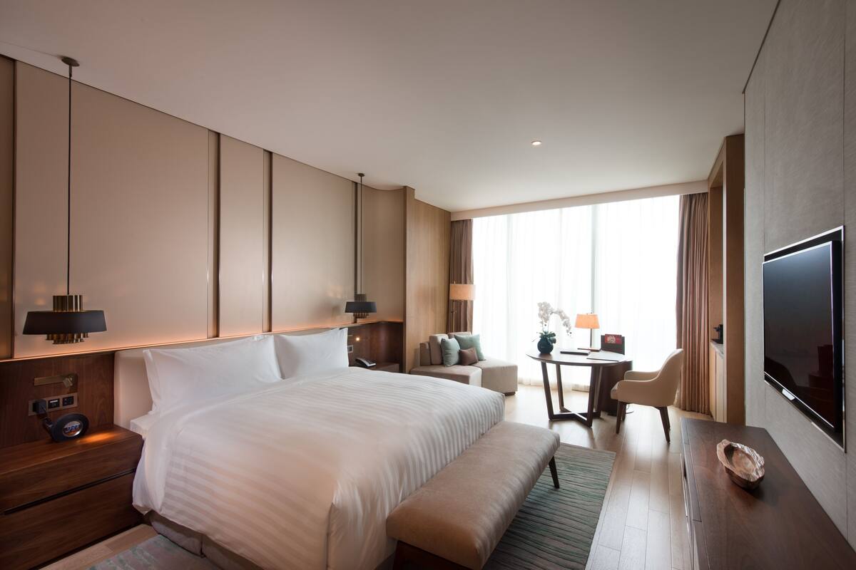 A hotel room with a large bed, bedside tables, a desk, a chair, a TV, and a window with curtains.