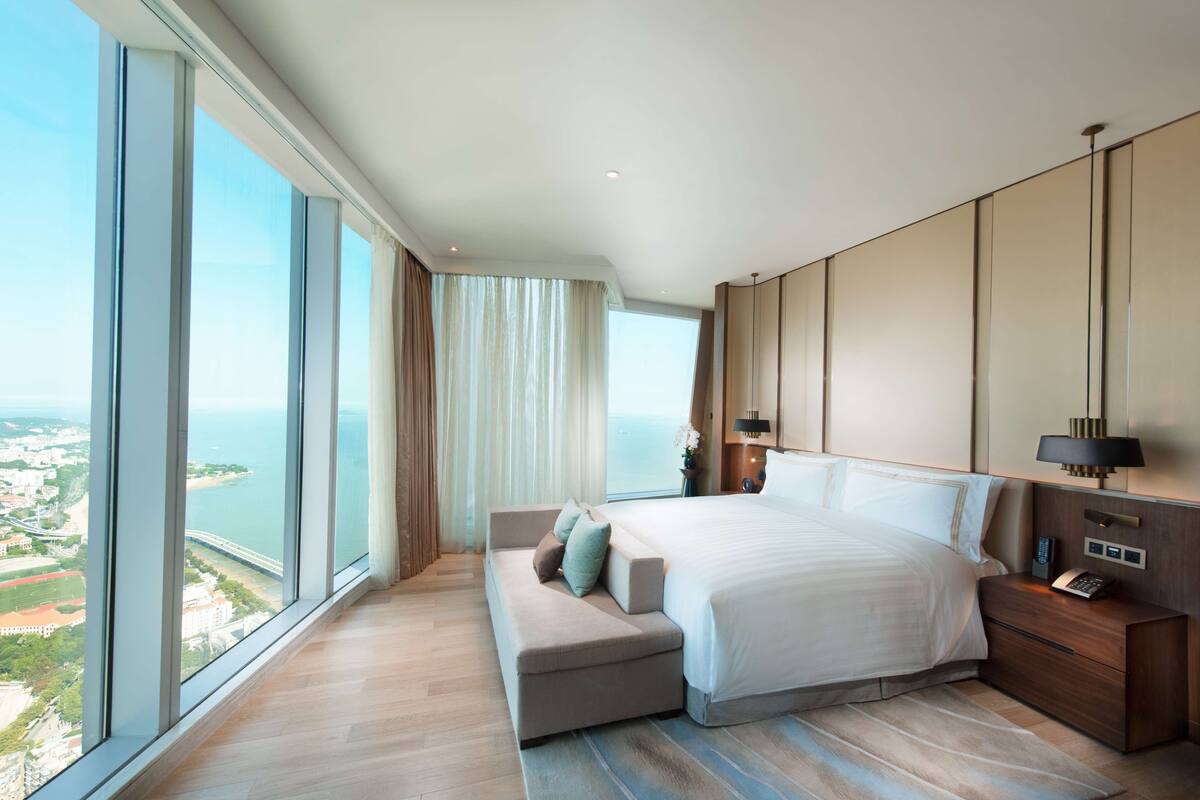 A modern bedroom with a large window, a bed, a nightstand, and a seating area.