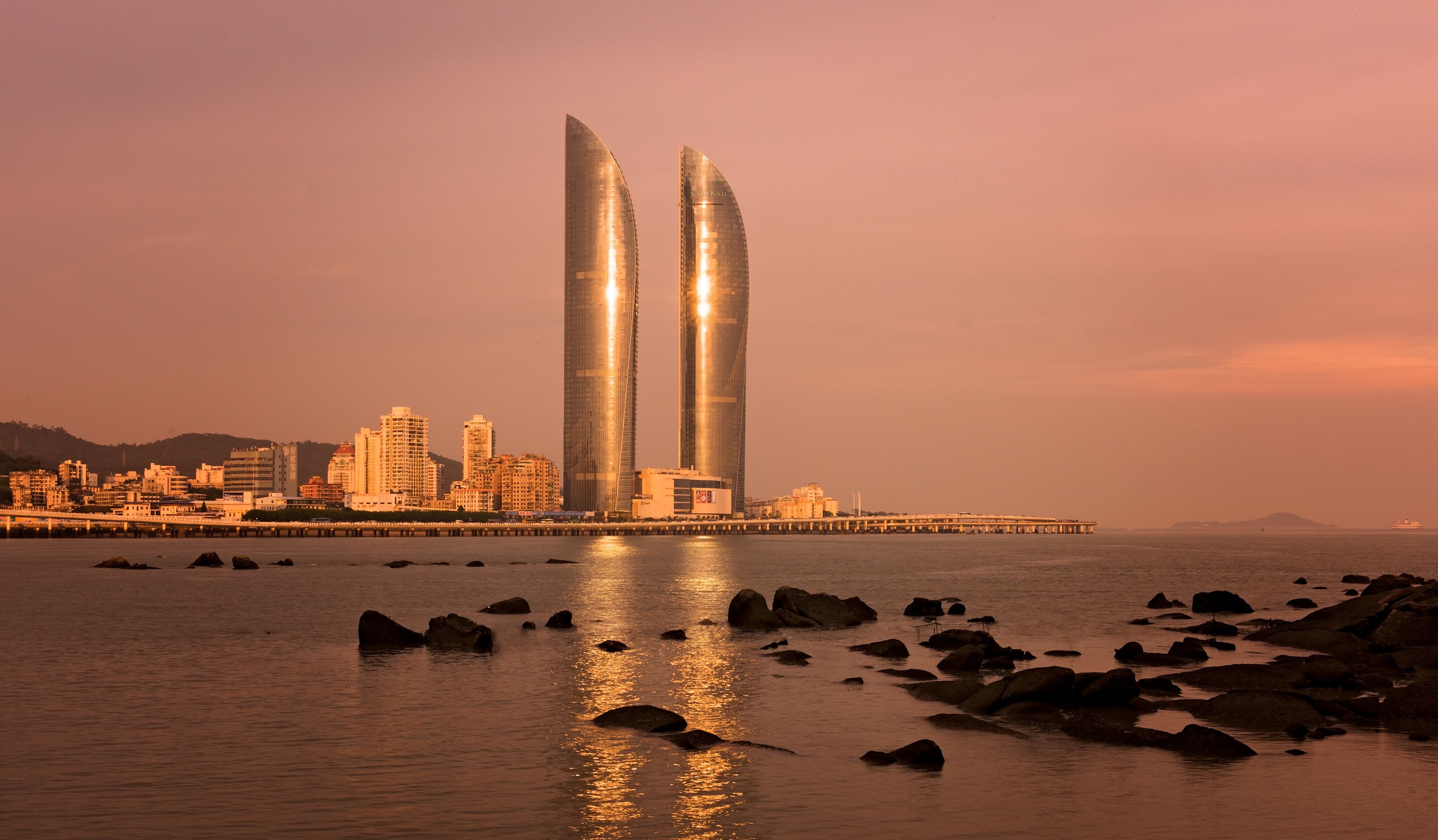 A cityscape with two prominent skyscrapers, a waterfront, and a pier extending into the water.