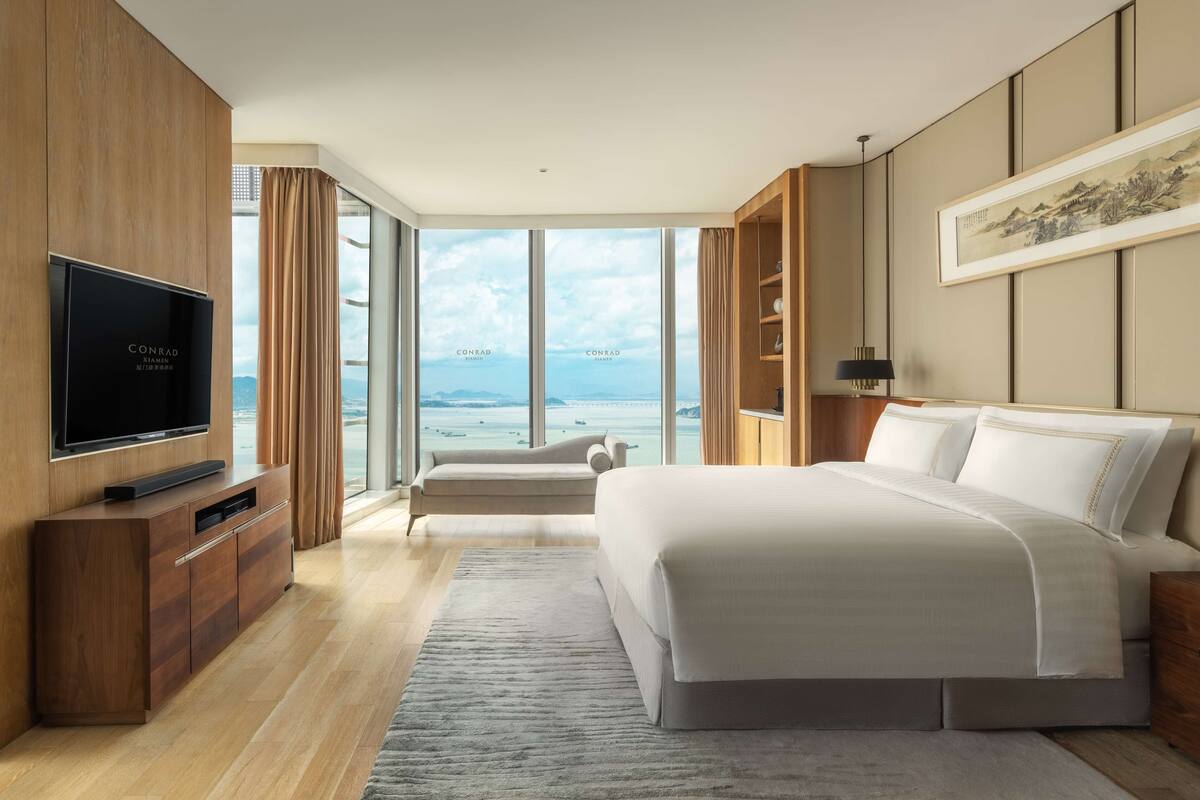 A modern hotel room with a large bed, a TV, a sofa, and a view of the sea.
