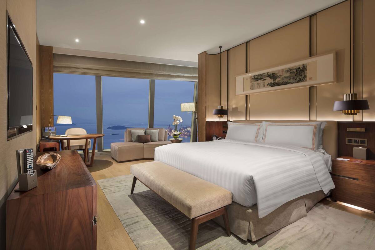 A modern hotel room with a large bed, a desk, a bench, and a view of the ocean.