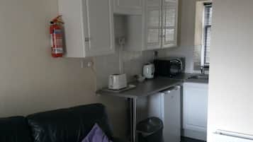 A compact living space with a kitchenette, a black leather sofa with a purple pillow, a white cabinet, a microwave, a toaster, and a fire extinguisher on the wall.