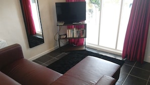 A living room with a brown sofa, a flat-screen TV on a stand, a bookshelf, and a sliding glass door with red curtains.