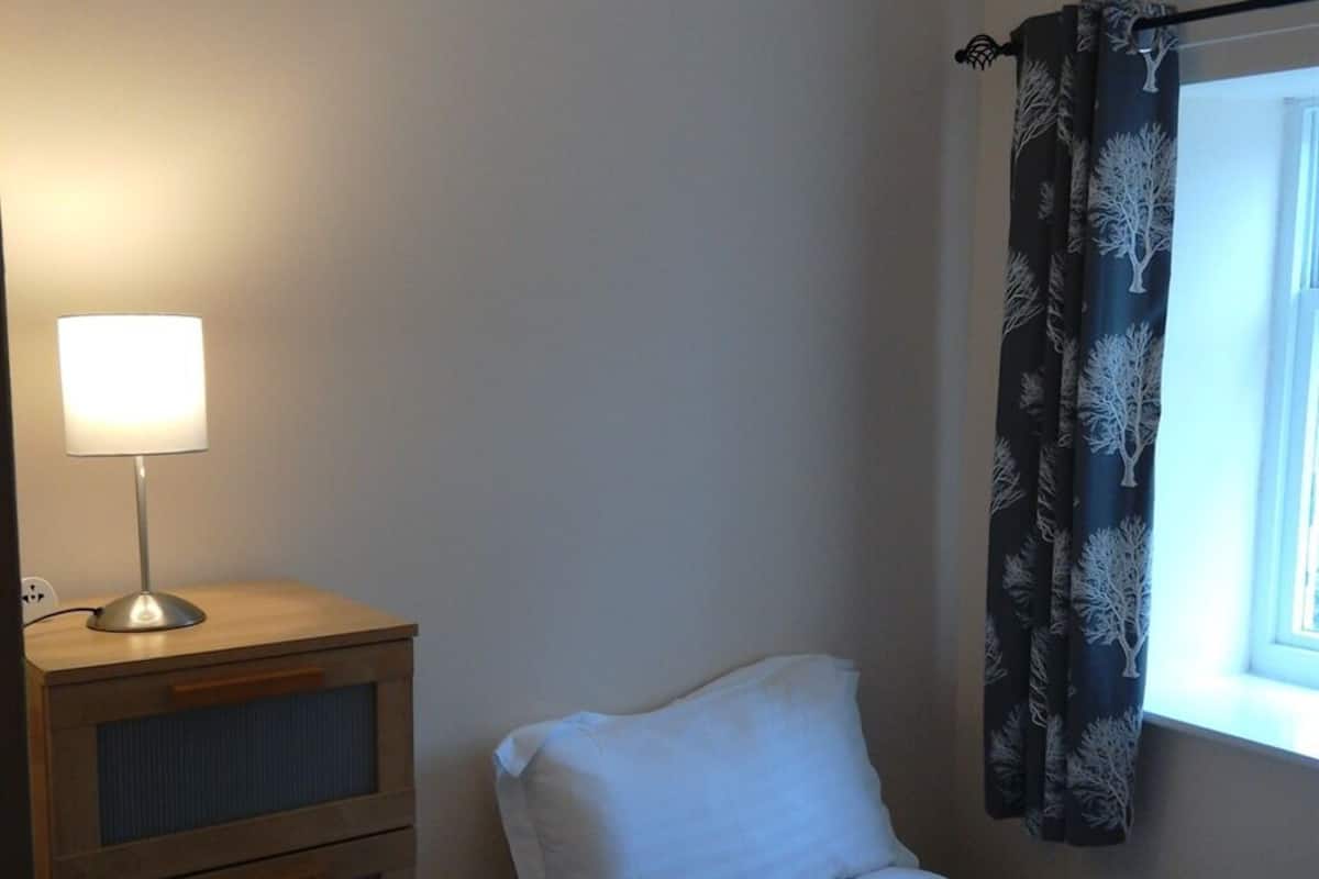 A small bedroom with a single bed, a bedside table with a lamp, and a window with curtains.