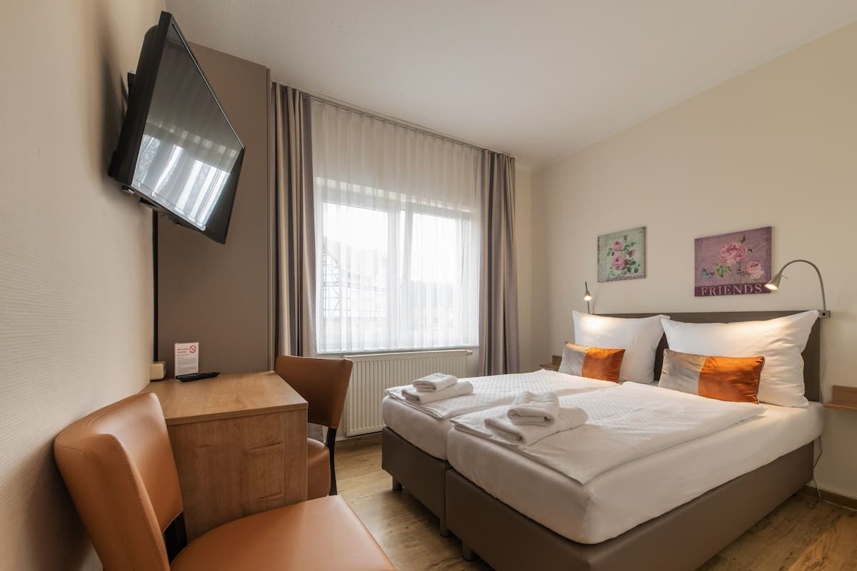 A hotel room with a bed, a desk, a chair, a TV mounted on the wall, and a window with curtains.
